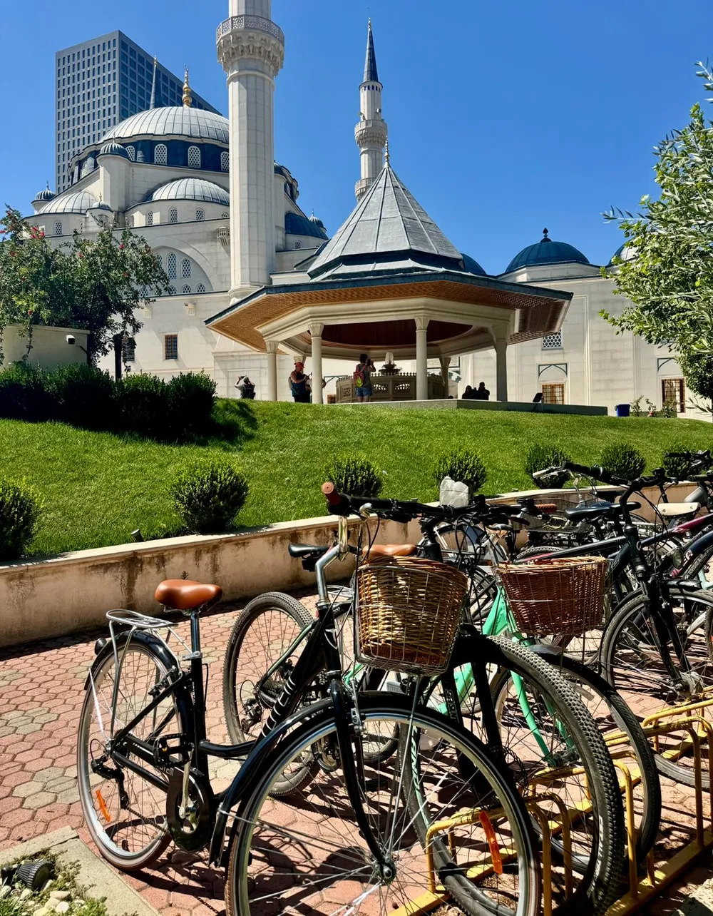 A bicycle with a wicker basket parked on a grassy lawn with the Namazgah Mosque of Tirana in the background
