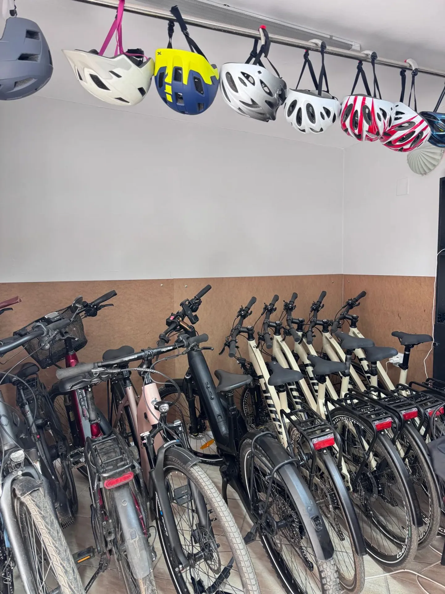 En række parkerede cykler med farverige hjelme hængende på et stativ ovenover i værkstedet